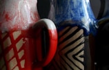 Two ceramic vases with intricate designs. The vase on the left features red and white marbled patterns with a prominent geometric grid, while the right vase showcases blue and cream zigzag motifs over a dark brown base.