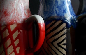 Two ceramic vases with intricate designs. The vase on the left features red and white marbled patterns with a prominent geometric grid, while the right vase showcases blue and cream zigzag motifs over a dark brown base.