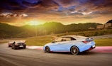 A group of sleek, modern cars race along a curving track with a backdrop of a stunning sunset over mountainous terrain. The golden sky casts warm hues across the landscape, adding an adventurous and dynamic atmosphere.