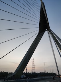 A close-up view of a cable-stayed bridge structure with multiple tension cables extending from the central support pylon, which has a triangular shape. In the background, there is an electricity pylon and a few street lights along the road beneath the bridge. The sky is mostly clear with a slight gradient, indicating either dawn or dusk.