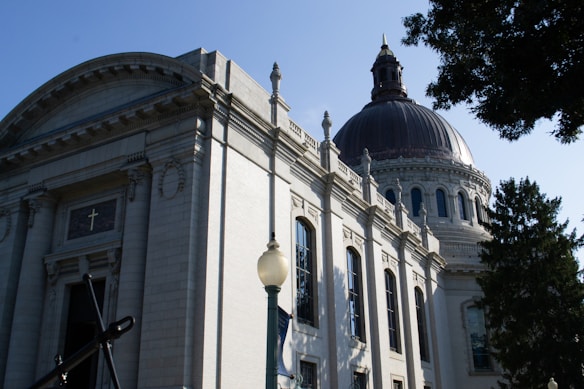 A large, ornate building with classical architecture, featuring a prominent dome and detailed stonework. The structure includes arched windows and decorative columns. A cross is visible above a doorway, suggesting a religious affiliation. Trees partially frame the scene, and there's a lamppost in the foreground.