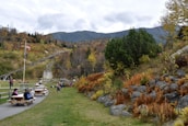 A scenic outdoor area with picnic tables and people enjoying leisure activities. A path runs through the greenery, flanked by rocky hills and trees starting to turn color in the fall. Flags are waving on poles, and a gentle slope leads up to a mountainous backdrop under an overcast sky.