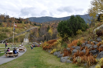 A scenic outdoor area with picnic tables and people enjoying leisure activities. A path runs through the greenery, flanked by rocky hills and trees starting to turn color in the fall. Flags are waving on poles, and a gentle slope leads up to a mountainous backdrop under an overcast sky.