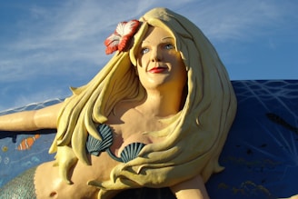 A large statue of a mermaid with long, flowing blonde hair adorned with a red and white flower. She has a serene expression and is depicted with a seashell bikini top. The background features an oceanic theme with fish and water details.