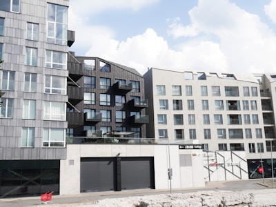 Modern apartment buildings with geometric designs and large windows dominate the scene. A prominent red shopping cart is placed in front of the structures, adding a splash of color to the predominantly grey facade. The sky is partly cloudy, creating a bright atmosphere.