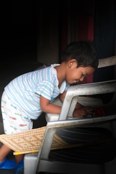A small child is playing with objects while leaning against a chair. The child is wearing a striped shirt and patterned shorts, and appears focused and engaged in play. A woven mat and other chairs are also visible in the scene.