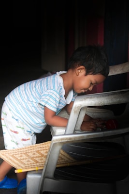 A small child is playing with objects while leaning against a chair. The child is wearing a striped shirt and patterned shorts, and appears focused and engaged in play. A woven mat and other chairs are also visible in the scene.
