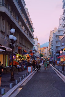 A vibrant urban street scene features a mix of people walking and interacting, lined with tall buildings on either side. The street is bustling with open shops, cafes, and street lamps. There is a relaxed and lively atmosphere, enhanced by the soft lighting at dusk.