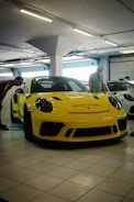 A bright yellow safety car is parked inside a garage with tiled floors. Two men are nearby, one examining the car and another standing beside it. The setting appears to be a car showroom or a display area with white overhead lighting and visible garage doors in the background.
