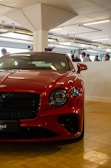 A luxurious red sports car is parked indoors on a wooden floor. The car's design features sharp lines and a sleek, polished finish. Several people in casual clothing are visible in the background, appearing to engage in conversation. The indoor setting is well-lit with fluorescent lights and a concrete structural column is present.