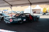 A custom racing car under a tent in a parking area, adorned with various sponsor logos including NGK and Liqui Moly. There is a person working inside the car, and nearby, there are other vehicles and tents with equipment. The area appears to be part of a racing event with noticeable branding and other automotive-related activities.