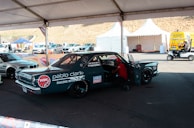 A custom racing car under a tent in a parking area, adorned with various sponsor logos including NGK and Liqui Moly. There is a person working inside the car, and nearby, there are other vehicles and tents with equipment. The area appears to be part of a racing event with noticeable branding and other automotive-related activities.