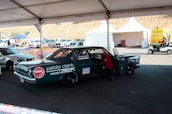 A custom racing car under a tent in a parking area, adorned with various sponsor logos including NGK and Liqui Moly. There is a person working inside the car, and nearby, there are other vehicles and tents with equipment. The area appears to be part of a racing event with noticeable branding and other automotive-related activities.