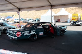 A custom racing car under a tent in a parking area, adorned with various sponsor logos including NGK and Liqui Moly. There is a person working inside the car, and nearby, there are other vehicles and tents with equipment. The area appears to be part of a racing event with noticeable branding and other automotive-related activities.