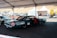 A custom racing car under a tent in a parking area, adorned with various sponsor logos including NGK and Liqui Moly. There is a person working inside the car, and nearby, there are other vehicles and tents with equipment. The area appears to be part of a racing event with noticeable branding and other automotive-related activities.