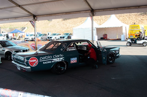 A custom racing car under a tent in a parking area, adorned with various sponsor logos including NGK and Liqui Moly. There is a person working inside the car, and nearby, there are other vehicles and tents with equipment. The area appears to be part of a racing event with noticeable branding and other automotive-related activities.