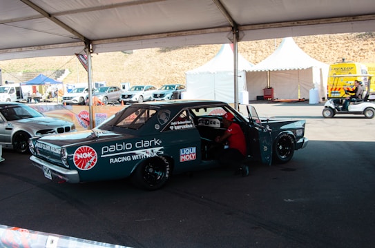 A custom racing car under a tent in a parking area, adorned with various sponsor logos including NGK and Liqui Moly. There is a person working inside the car, and nearby, there are other vehicles and tents with equipment. The area appears to be part of a racing event with noticeable branding and other automotive-related activities.