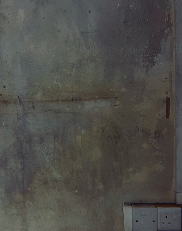 A textured wall with a rough, aged appearance featuring green and gray tones. The bottom right corner shows electrical outlets embedded into the wall, providing a modern contrast to the otherwise worn surface.