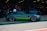 A sleek, high-performance sports car with a dark matte finish and bright green accents is parked in a garage area. Several people are standing nearby, some wearing lanyards, suggesting a car event or exhibition. The car has modern, aerodynamic features and large, stylish alloy wheels. The garage is dimly lit, adding a focused ambiance to the scene.