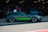 A sleek, high-performance sports car with a dark matte finish and bright green accents is parked in a garage area. Several people are standing nearby, some wearing lanyards, suggesting a car event or exhibition. The car has modern, aerodynamic features and large, stylish alloy wheels. The garage is dimly lit, adding a focused ambiance to the scene.