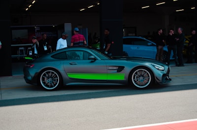 A sleek, high-performance sports car with a dark matte finish and bright green accents is parked in a garage area. Several people are standing nearby, some wearing lanyards, suggesting a car event or exhibition. The car has modern, aerodynamic features and large, stylish alloy wheels. The garage is dimly lit, adding a focused ambiance to the scene.