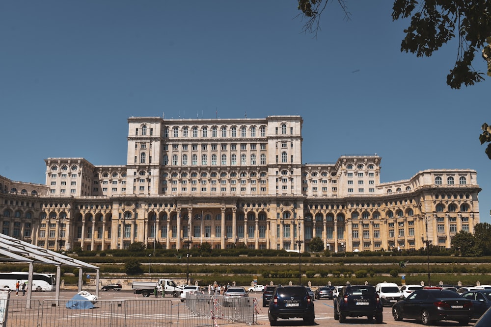 un grande edificio con auto parcheggiate di fronte ad esso con il Palazzo del Parlamento sullo sfondo