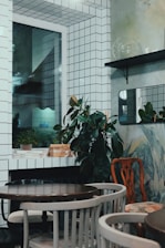 The image depicts a small, stylishly decorated room with white tiled walls and a window. There are potted plants placed on shelves and surfaces, adding a touch of greenery. The room is furnished with a few round wooden tables surrounded by a mix of chairs. There are minimalistic design elements with a mix of modern and vintage furniture.