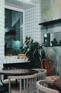 The image depicts a small, stylishly decorated room with white tiled walls and a window. There are potted plants placed on shelves and surfaces, adding a touch of greenery. The room is furnished with a few round wooden tables surrounded by a mix of chairs. There are minimalistic design elements with a mix of modern and vintage furniture.
