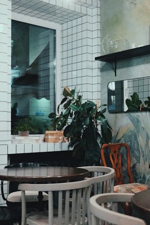 The image depicts a small, stylishly decorated room with white tiled walls and a window. There are potted plants placed on shelves and surfaces, adding a touch of greenery. The room is furnished with a few round wooden tables surrounded by a mix of chairs. There are minimalistic design elements with a mix of modern and vintage furniture.