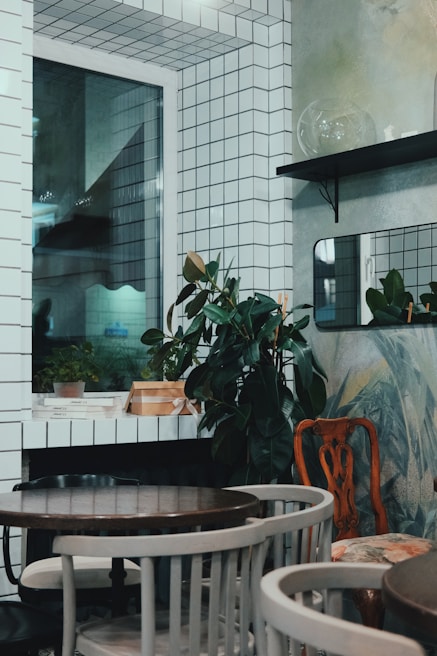 The image depicts a small, stylishly decorated room with white tiled walls and a window. There are potted plants placed on shelves and surfaces, adding a touch of greenery. The room is furnished with a few round wooden tables surrounded by a mix of chairs. There are minimalistic design elements with a mix of modern and vintage furniture.