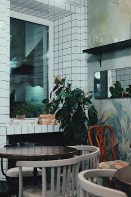 The image depicts a small, stylishly decorated room with white tiled walls and a window. There are potted plants placed on shelves and surfaces, adding a touch of greenery. The room is furnished with a few round wooden tables surrounded by a mix of chairs. There are minimalistic design elements with a mix of modern and vintage furniture.
