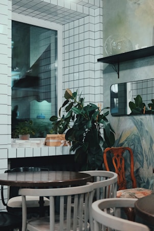 The image depicts a small, stylishly decorated room with white tiled walls and a window. There are potted plants placed on shelves and surfaces, adding a touch of greenery. The room is furnished with a few round wooden tables surrounded by a mix of chairs. There are minimalistic design elements with a mix of modern and vintage furniture.