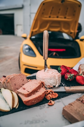 A picnic setup with slices of bread, strawberries, pear slices, and cheese with a knife placed into it, arranged on a black serving board. In the background, a yellow sports car is parked with its hood open.