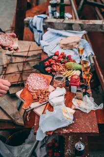 A rustic table is covered with an assortment of food and drink. There are slices of cured meat on a wooden board, alongside walnuts and a wedge of cheese. A variety of fruits, including strawberries and what appears to be an avocado, are scattered nearby. There are several small glasses filled with a golden liquid, likely a kind of liquor. A jar with red jam, a grater, and some bread are also present. An open package reveals more cheese, and a hand is holding a piece of bread with a slice of meat.