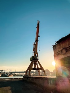 An industrial scene featuring a large crane situated near a dock. The crane is positioned prominently against a clear blue sky with the sun setting in the background, casting a warm yellowish glow. Surrounding the crane are various industrial structures and warehouses, some partially silhouetted by the sunlight.