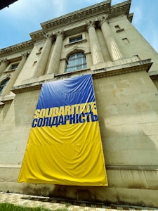 A large building with classical architectural features, including tall columns and decorative stonework. There is a prominent banner draped over the facade, featuring the word 'SOLIDARITATE' and its equivalent in another language, printed in blue on a yellow background.