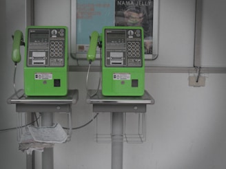 Two green public payphones are mounted on a wall. Below each phone, there are metal baskets for holding items, and one contains newspapers. A poster is visible in the background.