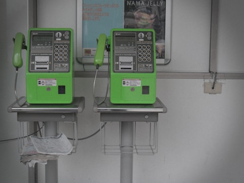 Two green public payphones are mounted on a wall. Below each phone, there are metal baskets for holding items, and one contains newspapers. A poster is visible in the background.