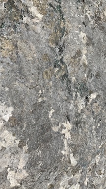 A textured surface with various shades of gray, white, and hints of beige resembling natural stone or rock. It features irregular patterns and speckled areas, giving it a rugged and natural appearance.