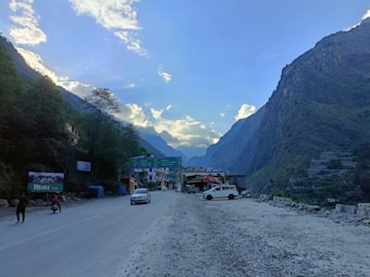 A scenic mountainous landscape with a road stretching into the distance, flanked by steep hills on both sides. There are a few vehicles parked on the roadside, and some buildings are visible near a large signboard. Trees and clouds are abundant, contributing to a serene atmosphere.