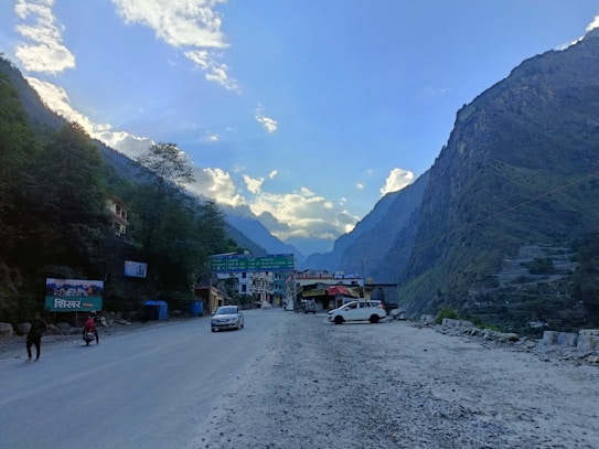 A scenic mountainous landscape with a road stretching into the distance, flanked by steep hills on both sides. There are a few vehicles parked on the roadside, and some buildings are visible near a large signboard. Trees and clouds are abundant, contributing to a serene atmosphere.