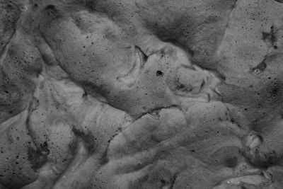 A textured surface with an organic appearance, possibly resembling foam or rock with various indentations and patterns. The surface has a monochromatic tone with visible pores and shadows.