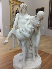 A marble sculpture depicting three figures in an emotional scene, with two adults holding a smaller, limp figure. The sculpture is placed on a circular base in a gallery setting, with framed artwork visible in the background.