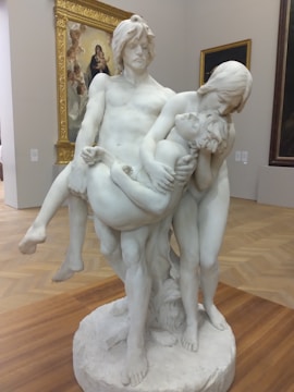 A marble sculpture depicting three figures in an emotional scene, with two adults holding a smaller, limp figure. The sculpture is placed on a circular base in a gallery setting, with framed artwork visible in the background.