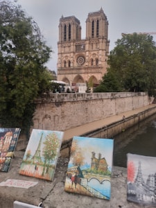A historic gothic cathedral stands majestically by a river, with its iconic two towers and intricate facade visible. In the foreground, several vibrant paintings are displayed on a stone ledge, depicting various parisian scenes including the Eiffel Tower.