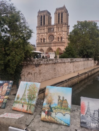 A historic gothic cathedral stands majestically by a river, with its iconic two towers and intricate facade visible. In the foreground, several vibrant paintings are displayed on a stone ledge, depicting various parisian scenes including the Eiffel Tower.