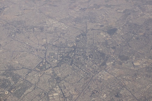 Aerial view of a densely populated urban area with a complex network of streets and buildings. The landscape is predominantly flat, showcasing a grid-like pattern of infrastructure and various clusters of structures, likely indicative of residential, commercial, and possibly some green areas.