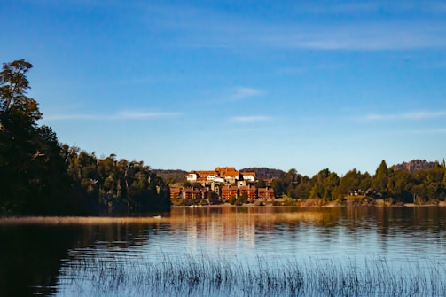 A serene landscape with a large, elegant building surrounded by lush greenery. The structure sits atop a hill bordered by a body of water, reflecting the blue sky and trees. The scene is peaceful, with tall grass visible at the water's edge and a backdrop of distant hills.