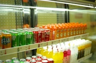 Rows of colorful beverage cans and bottles are neatly arranged on shelves inside a refrigerated display. The top shelf features green, red, and orange cans, while the bottom shelf holds yellow, pink, and white bottles.