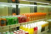 Rows of colorful beverage cans and bottles are neatly arranged on shelves inside a refrigerated display. The top shelf features green, red, and orange cans, while the bottom shelf holds yellow, pink, and white bottles.